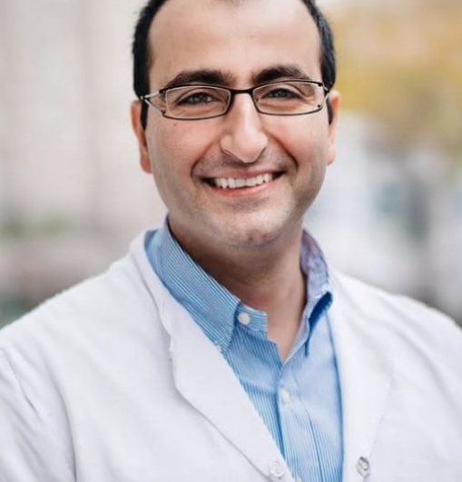 Dr. Michel Chraim in weisser Kleidung und einem hellblau-gestreiften Hemd sieht lachend nach vorne, er trägt eine dunkle Brille, man sieht Kopf und Oberkörper